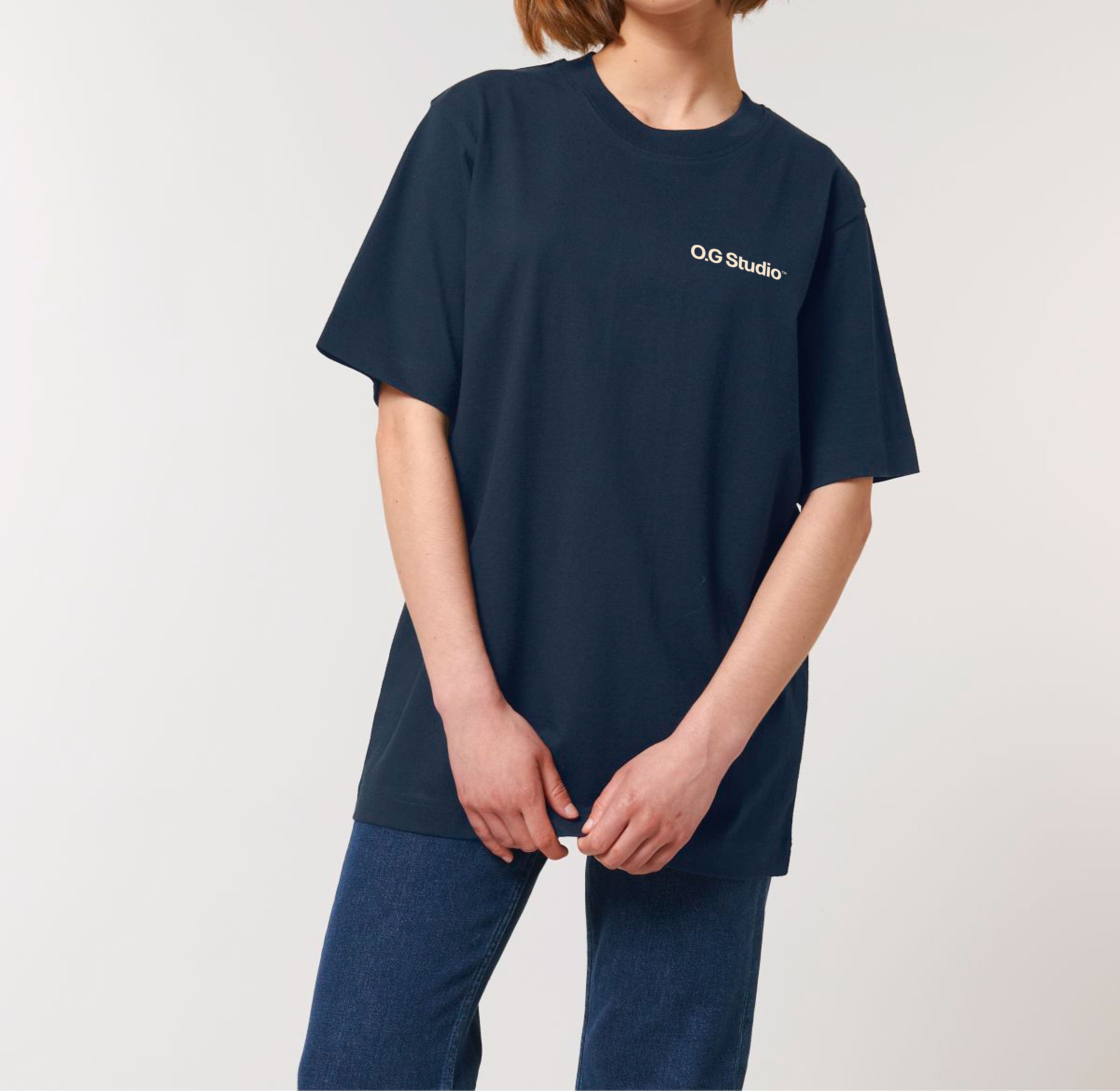 Model står oprejst iført mørkeblå T-shirt med hvidt “OG Studio” frontlogo, kombineret med mørkeblå jeans. Billedet fokuserer på T-shirtens pasform og minimalistiske design, der skaber et tidløst og moderne streetwear-udtryk. Den neutrale baggrund sikrer, at produktet er i centrum.