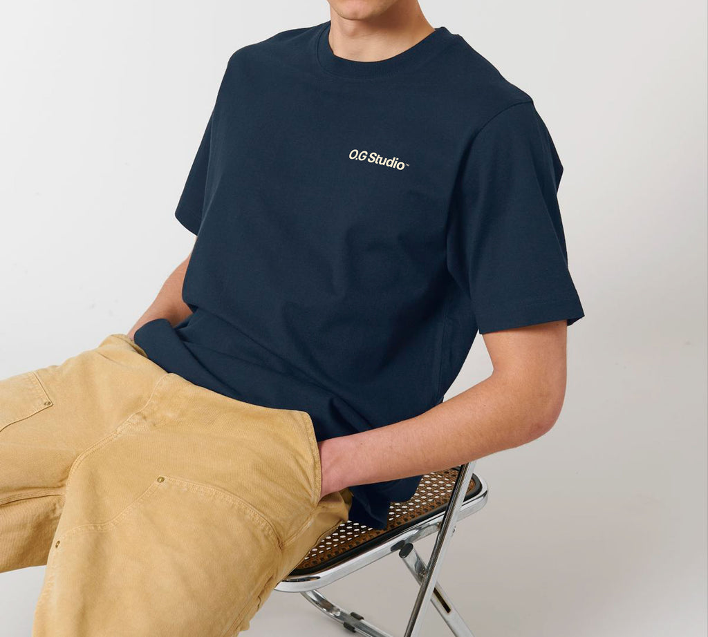 Person sidder afslappet på en stol iført mørkeblå T-shirt med lille hvidt "OG Studio" logo på brystet. T-shirten kombineres med lysebrune bukser, og posituren fremhæver det casual look og T-shirtens pasform. Den enkle styling og neutrale baggrund gør billedet velegnet til at præsentere produktets alsidighed og komfort.