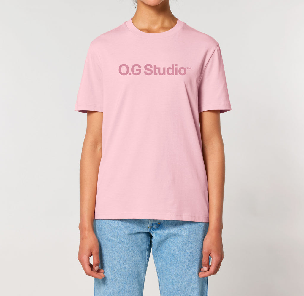Person iført lyserød T-shirt med mørkerødt “OG Studio” logo trykt på brystet. Modellen står oprejst med armene afslappet langs siden, iført lyseblå jeans. Den lyse baggrund fremhæver T-shirtens minimalistiske og moderne design, som kombinerer stilren æstetik med casual streetwear.